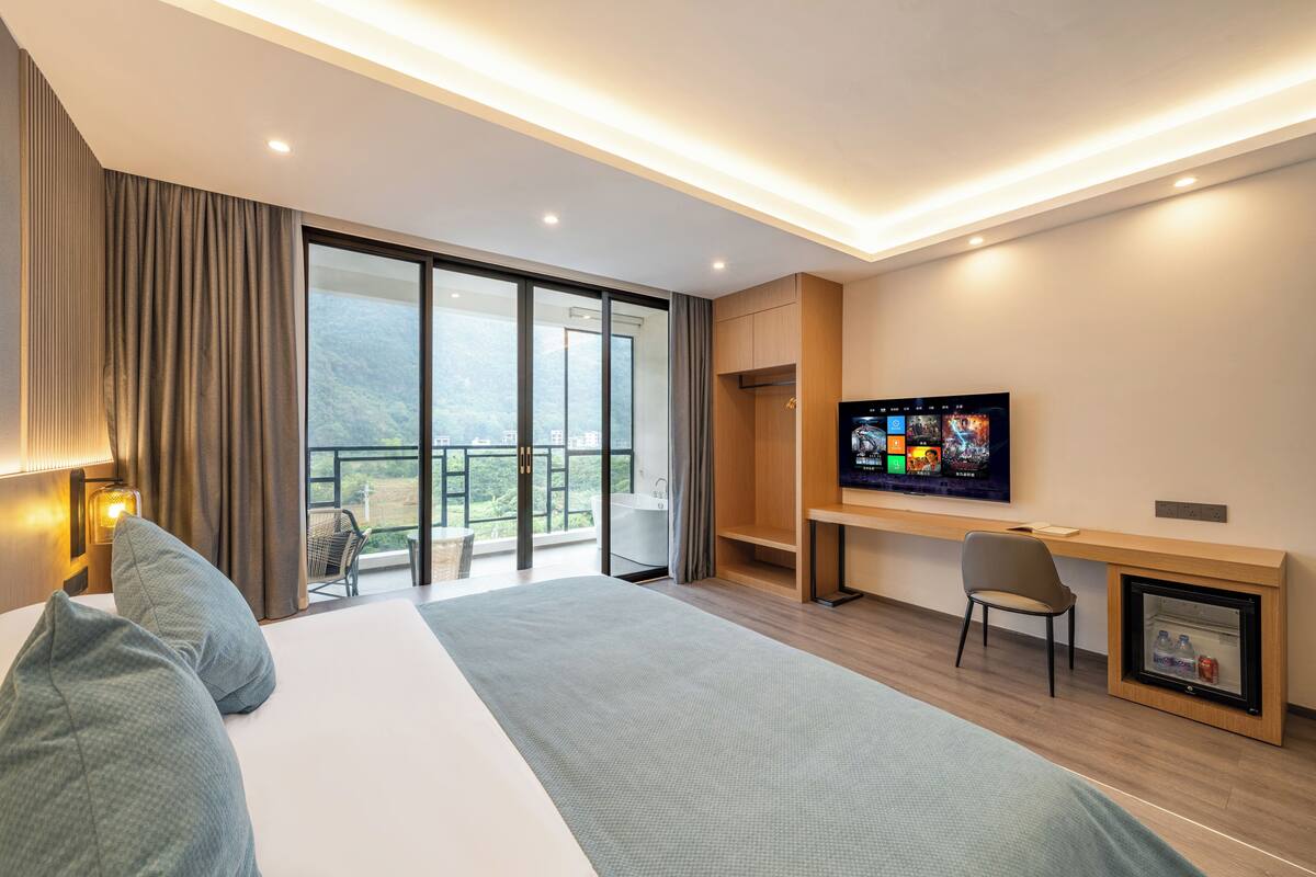 Quarto de hotel moderno com uma cama grande, uma escrivaninha, televisão e varanda com vista para a cidade.