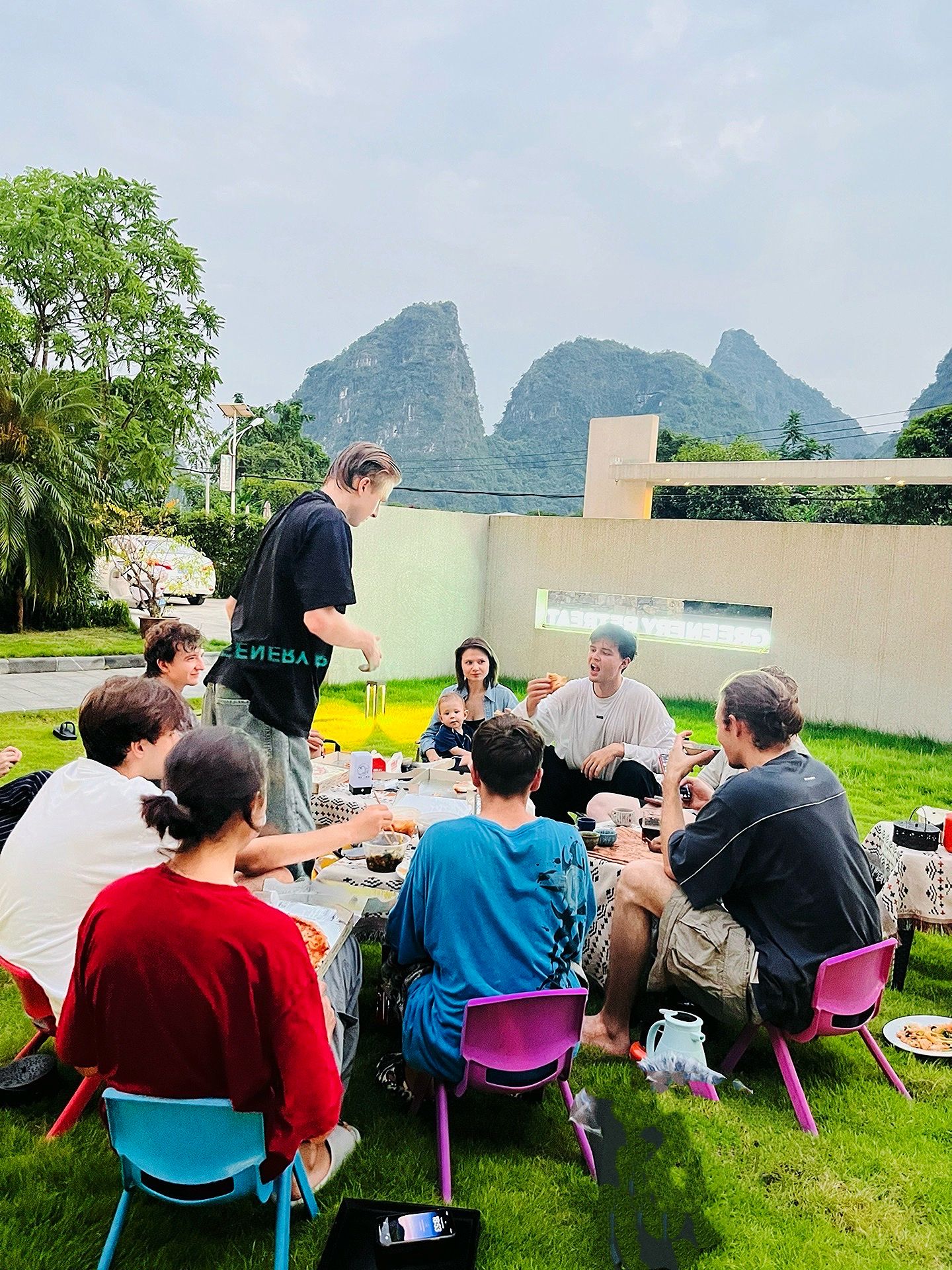 A group of people seated outdoors on grass, engaged in conversation and eating, with a mountainous landscape in the background.