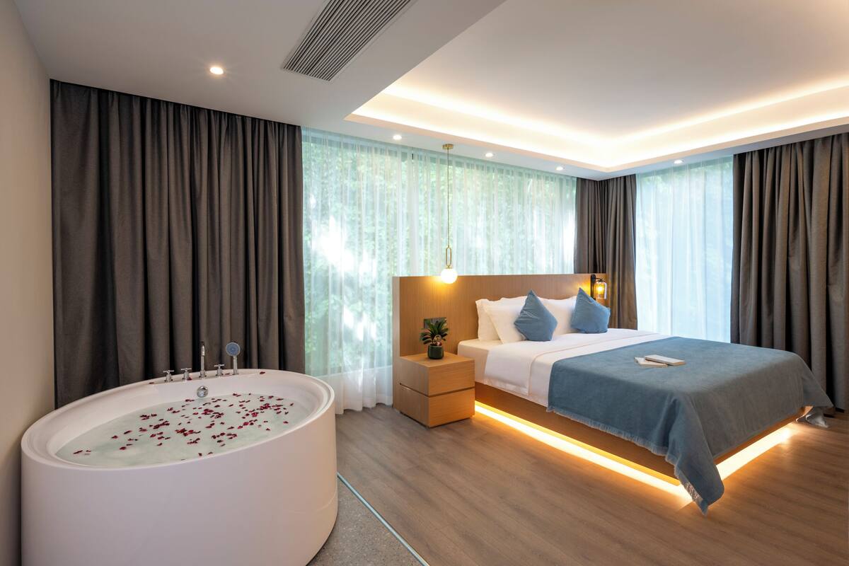 Quarto de hotel moderno com uma banheira grande, uma cama bem arrumada com manta azul e uma mesinha de cabeceira de madeira com uma planta.