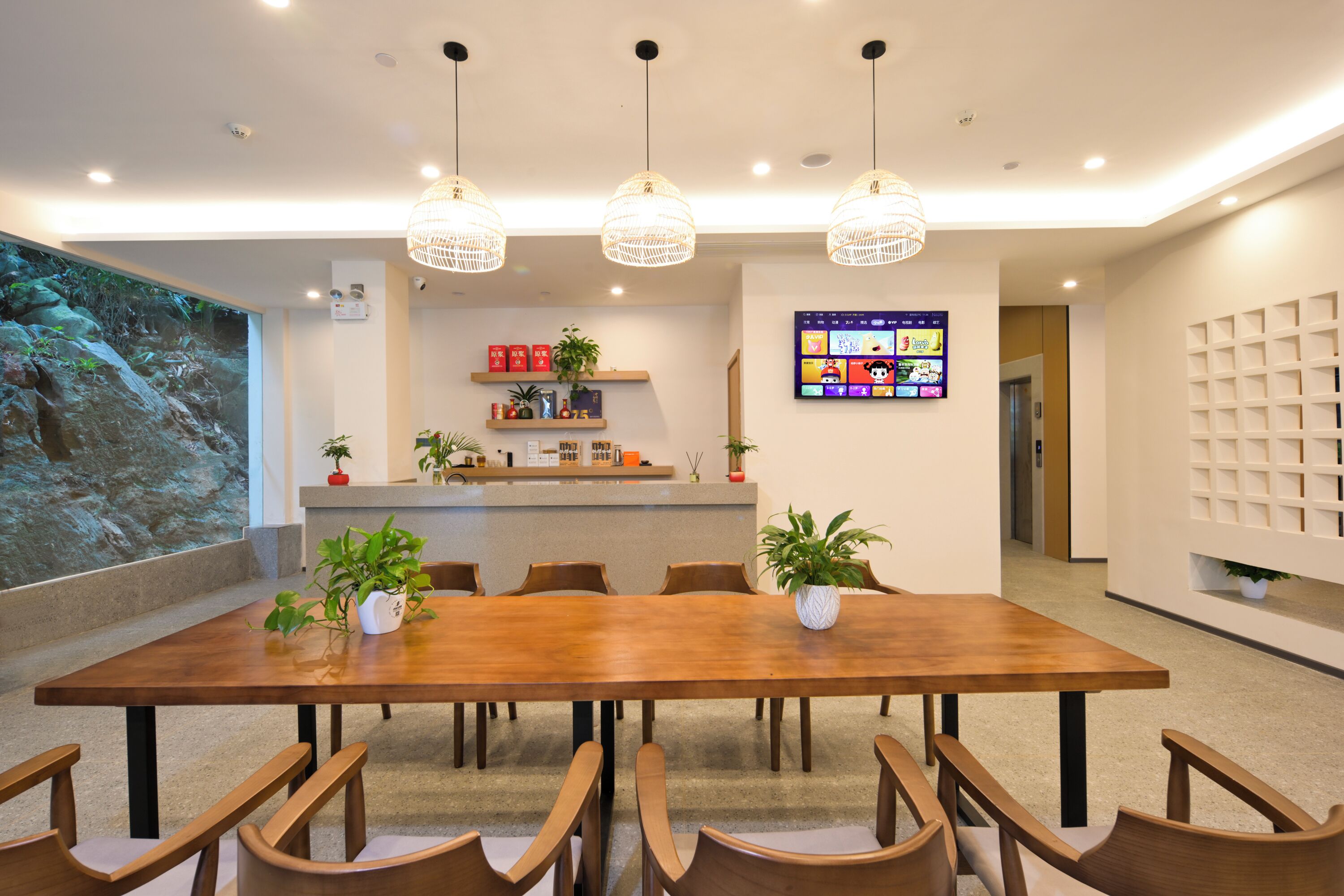 A modern conference room with a wooden table, chairs, pendant lights, a digital screen, and a wall-mounted shelf with plants.
