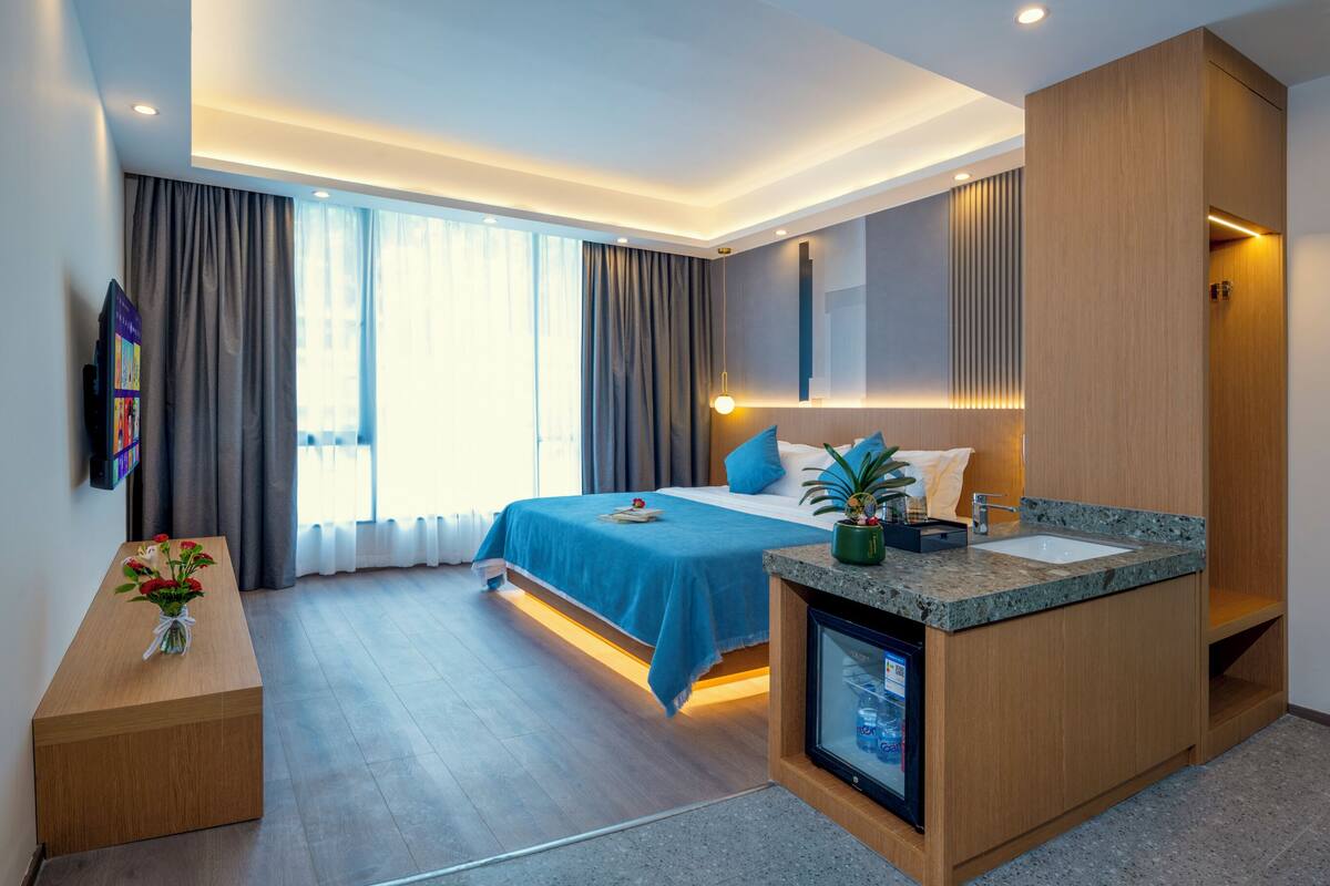 Quarto de hotel moderno com uma cama, TV integrada, mini-frigorífico e banheiro com pia e espelho.