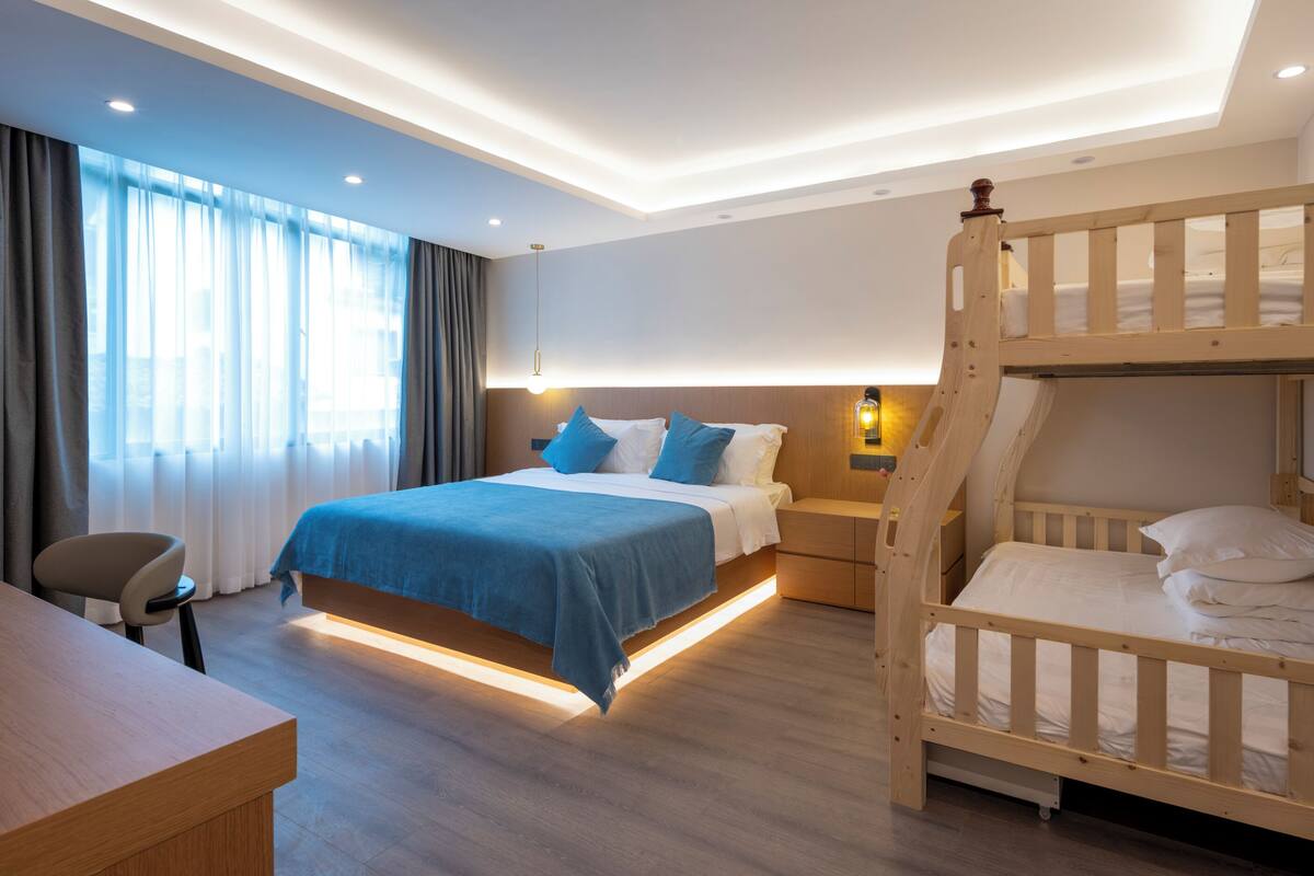 Quarto de hotel com uma cama beliche, uma escrivaninha, uma cadeira e uma janela grande com cortinas.
