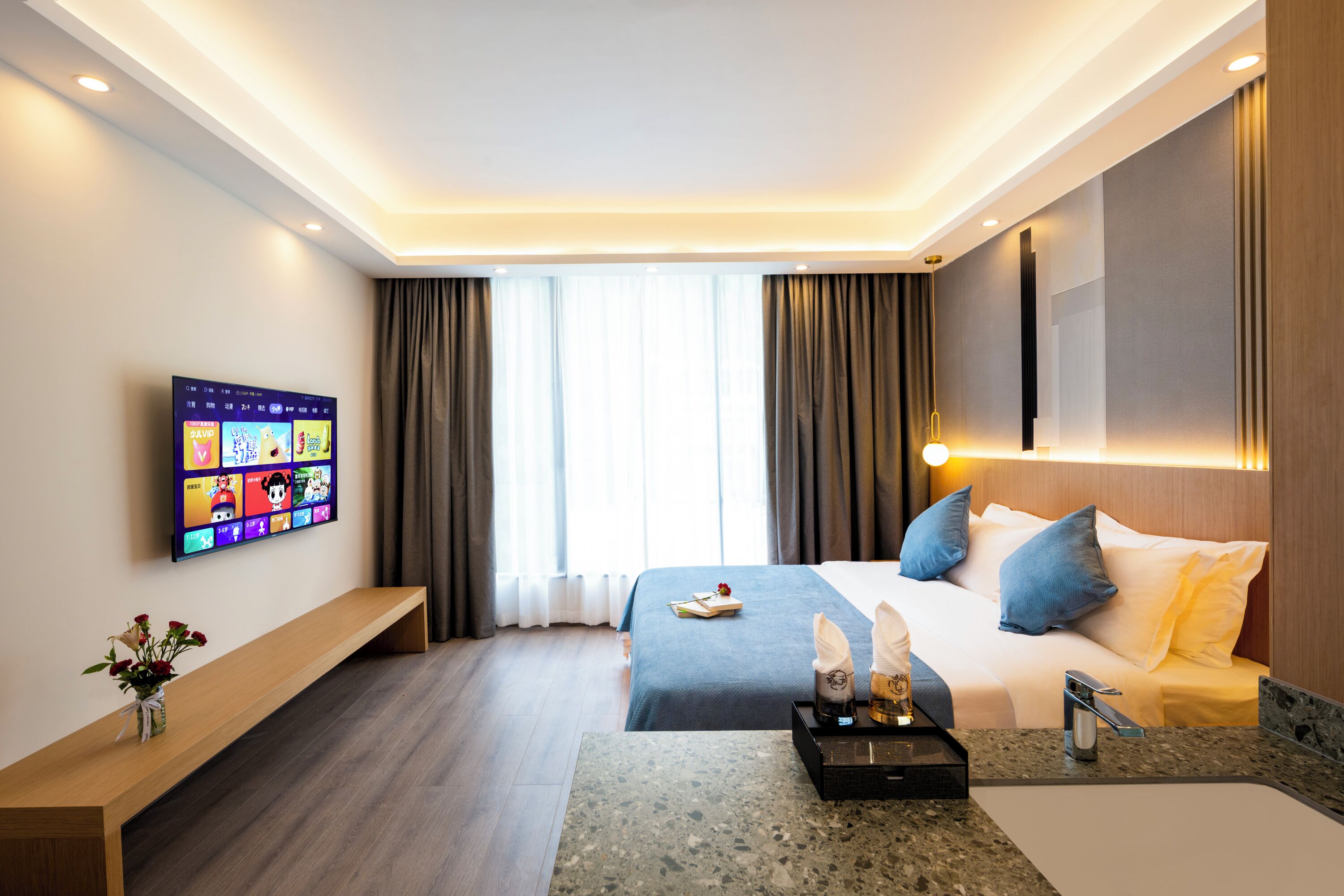 Une chambre d’hôtel moderne dotée d’un grand lit, d’un téléviseur à écran plat fixé au mur, d’un banc en bois et d’un plan de travail en marbre avec un évier.