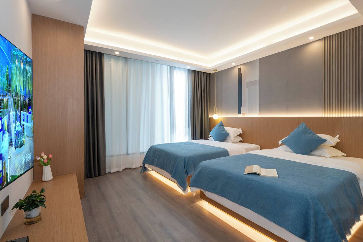 Quarto de hotel moderno com duas camas, televisão e uma janela grande com cortinas.