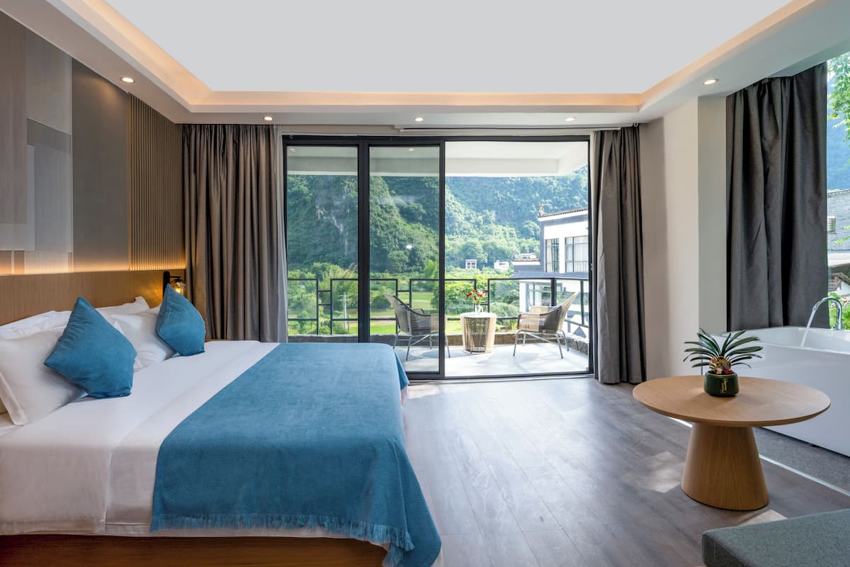 Quarto de hotel moderno com uma cama grande, uma mesa de madeira redonda e uma varanda com vista para áreas verdes e um edifício.
