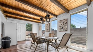 A dining area with a wooden ceiling, a table with chairs, and a view through large windows.