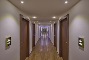 A long, well-lit hallway with wooden floors and doors, leading to a distant exit.
