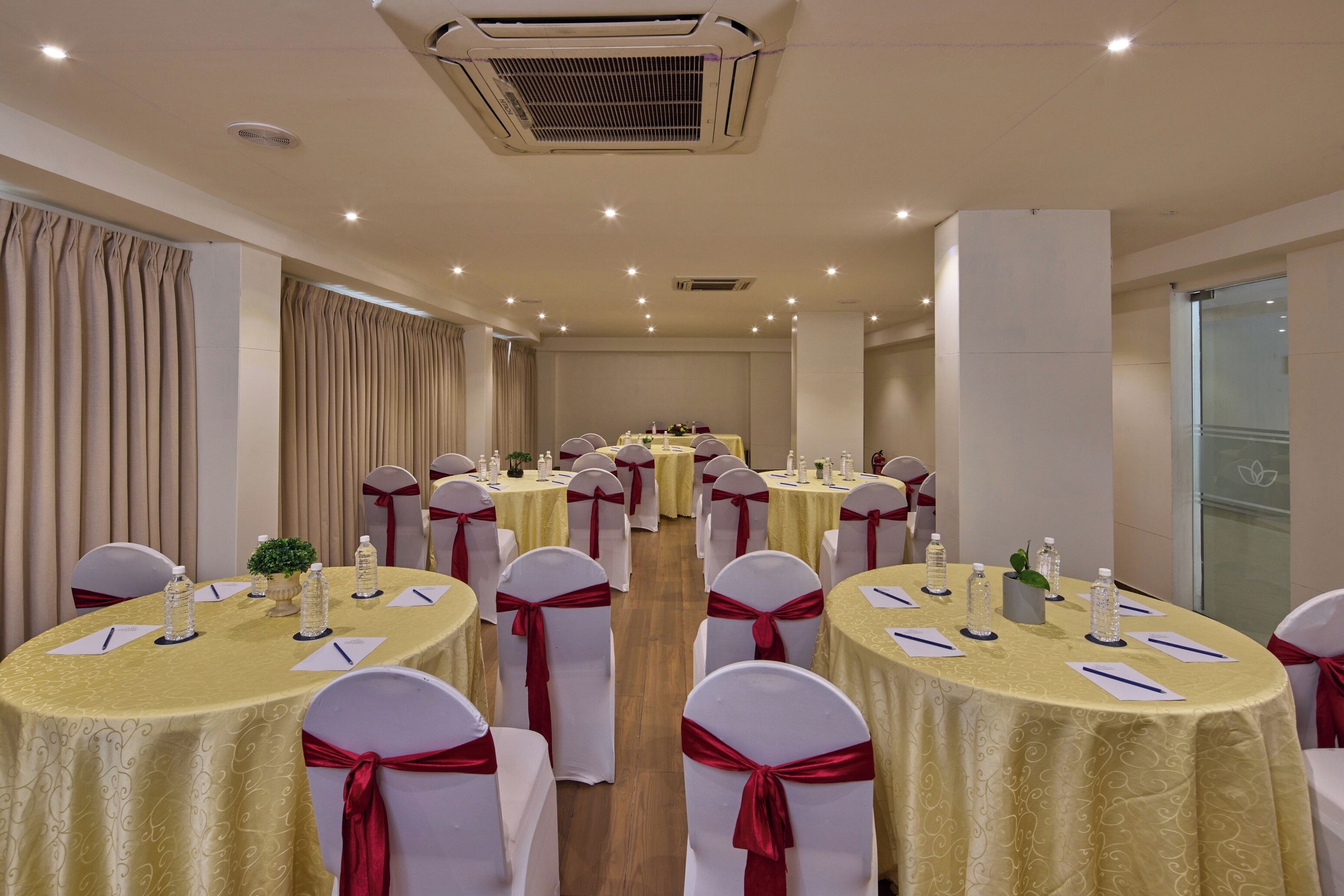 A conference room with round tables, white chairs, and red sashes. Tables are set with water bottles and notepads.