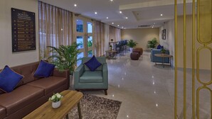 A hotel lobby with a brown leather sofa, teal armchair, and a wooden coffee table with a flower vase. A signboard lists room numbers and amenities. - Grand Continent Brookefield – A Sarovar Portico Affiliate Hotel (Bengaluru)