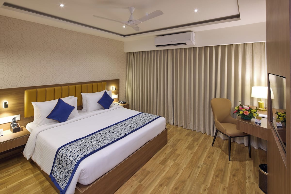 Quarto de hotel com cama, mesa de cabeceira, cadeira, escrivaninha e televisão.