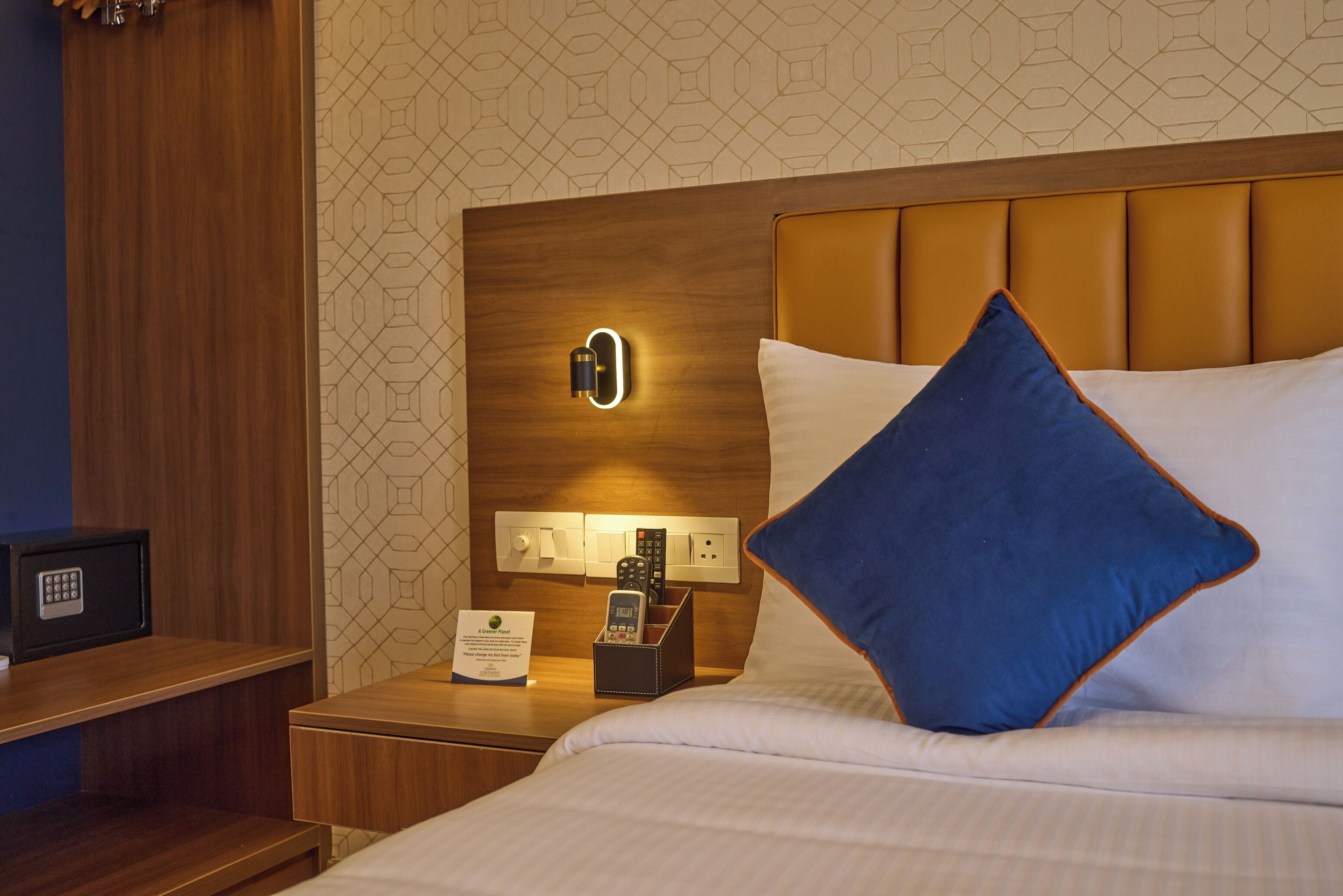 A hotel room with a wooden headboard, a bedside table with a phone and a sign, a blue pillow, and a patterned wall.