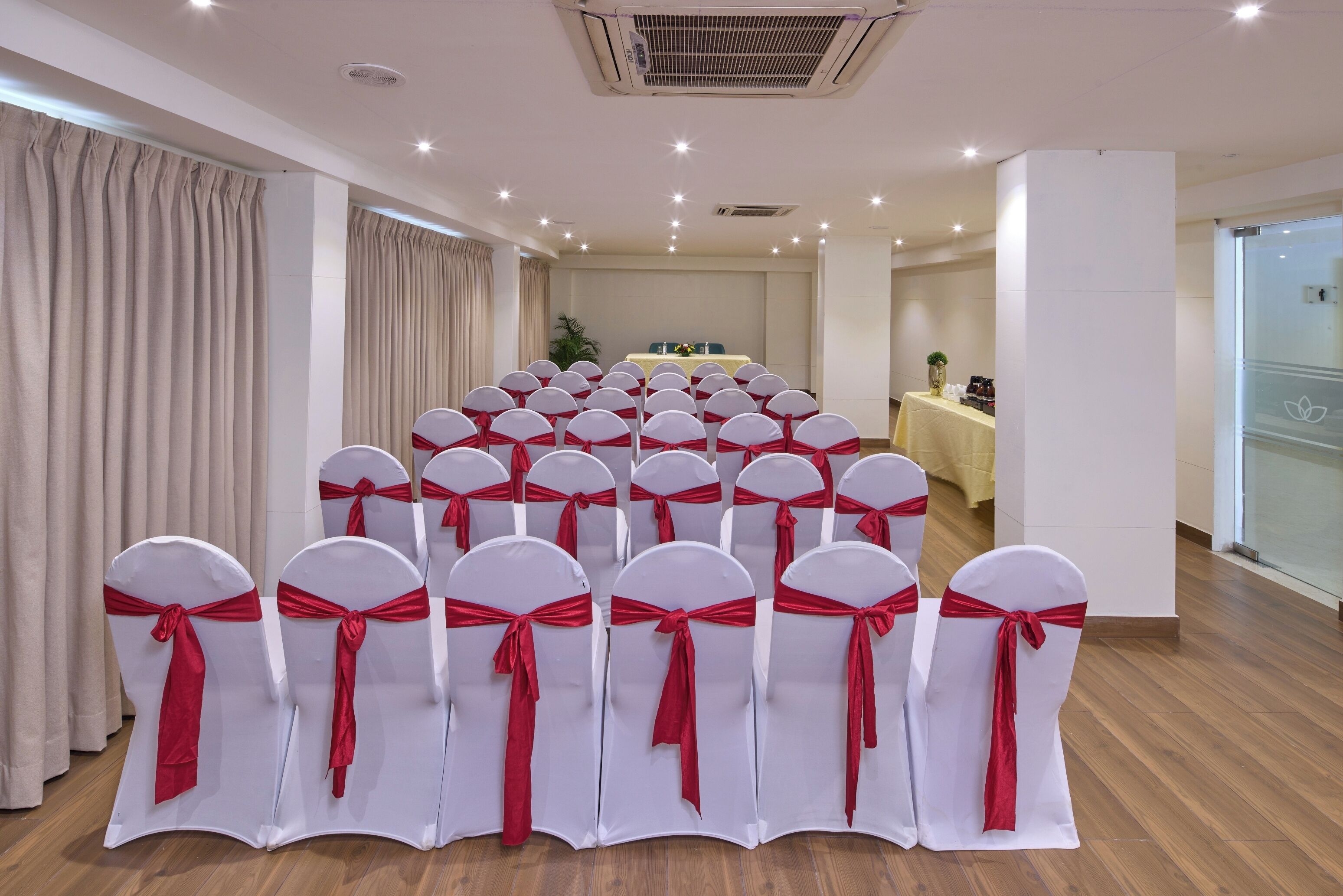 A conference room with rows of white chairs with red sashes, a table with floral arrangements, and a glass partition on one side.