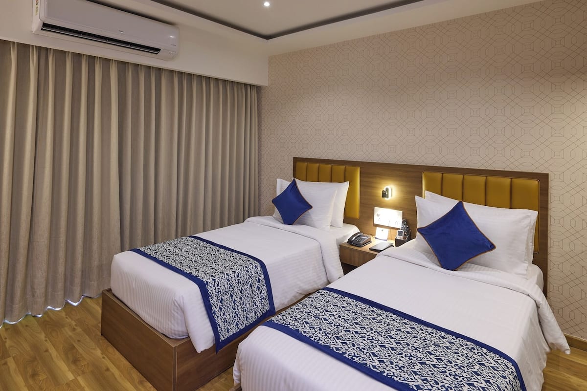 Quarto de hotel com duas camas, cabeceira de madeira, mesa de cabeceira com abajur e uma janela grande com cortinas.