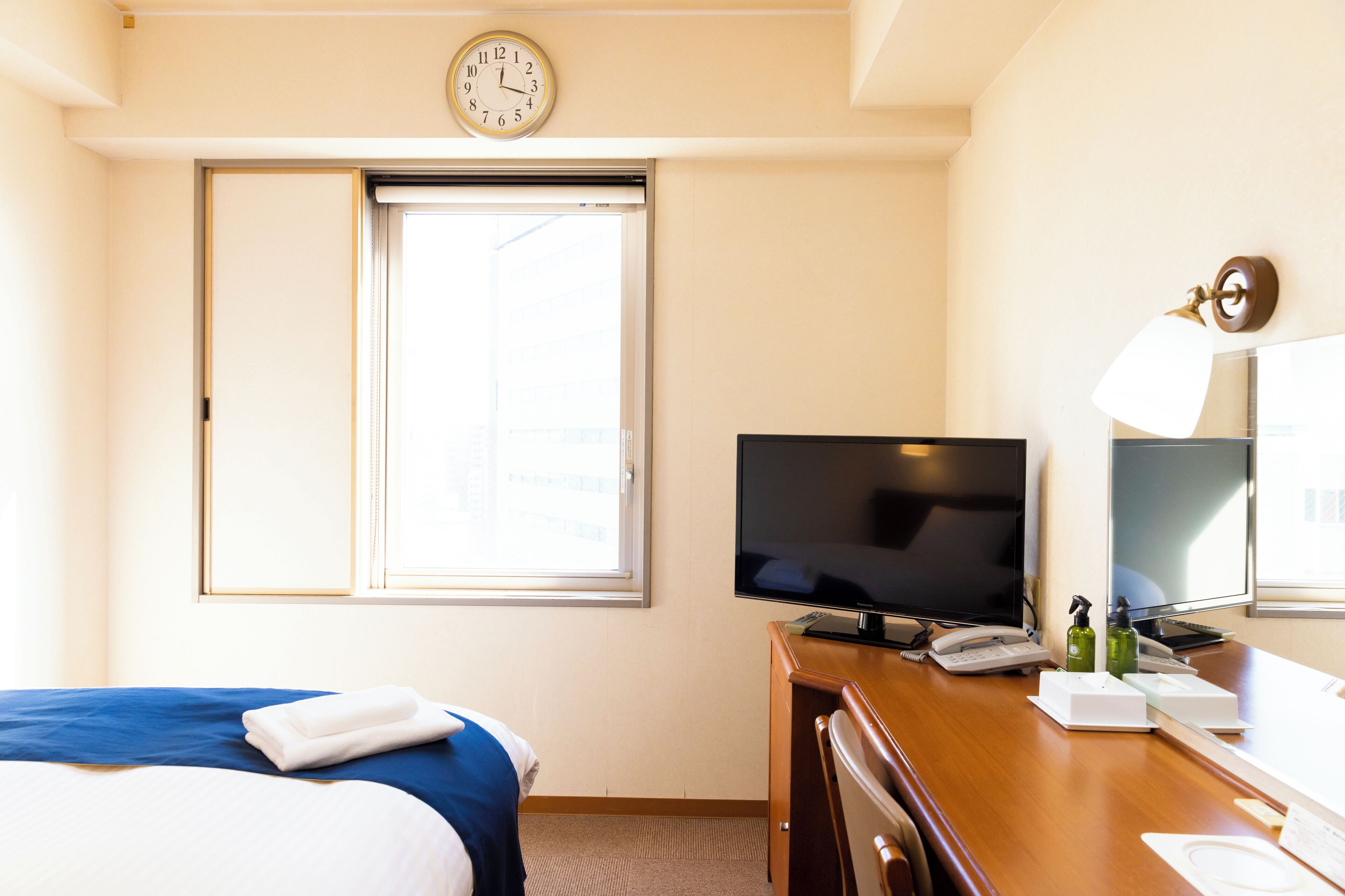 A hotel room with a bed, a desk with a television, a clock, and a window.