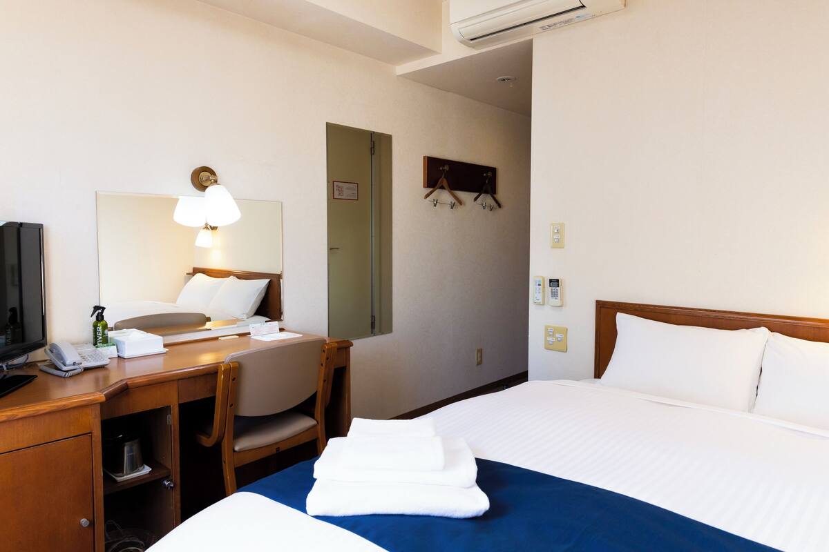 A hotel room with a bed, a desk with a telephone, a chair, a mirror, and a wall-mounted TV.