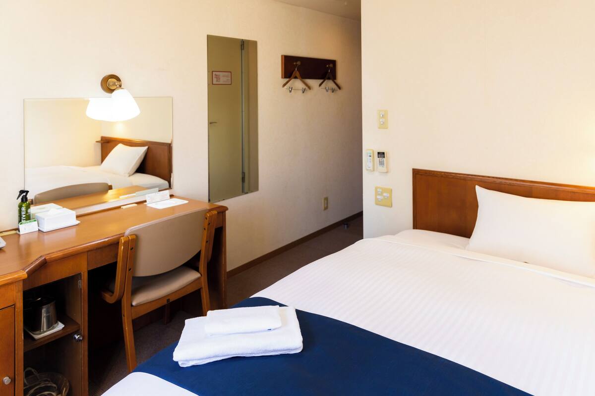 A hotel room with a bed, a desk, a chair, a mirror, a telephone, and a wall-mounted coat rack.