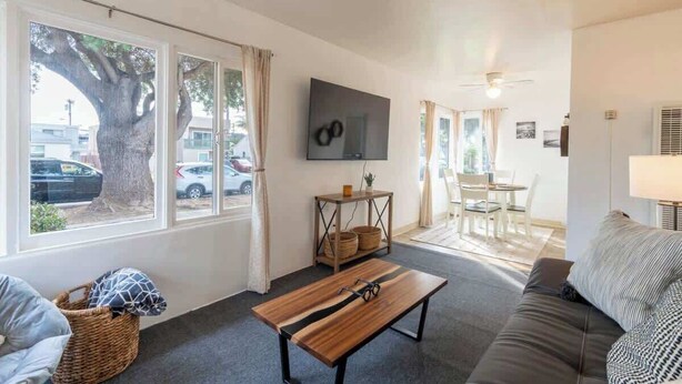 Un salón con sofá, una mesa de centro de madera, televisor montado en la pared, zona de comedor con mesa y sillas, y ventanas amplias con cortinas.
