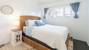 Un dormitorio con cama, mesita de noche, lámpara, ventana con cortinas y ventilador de techo.