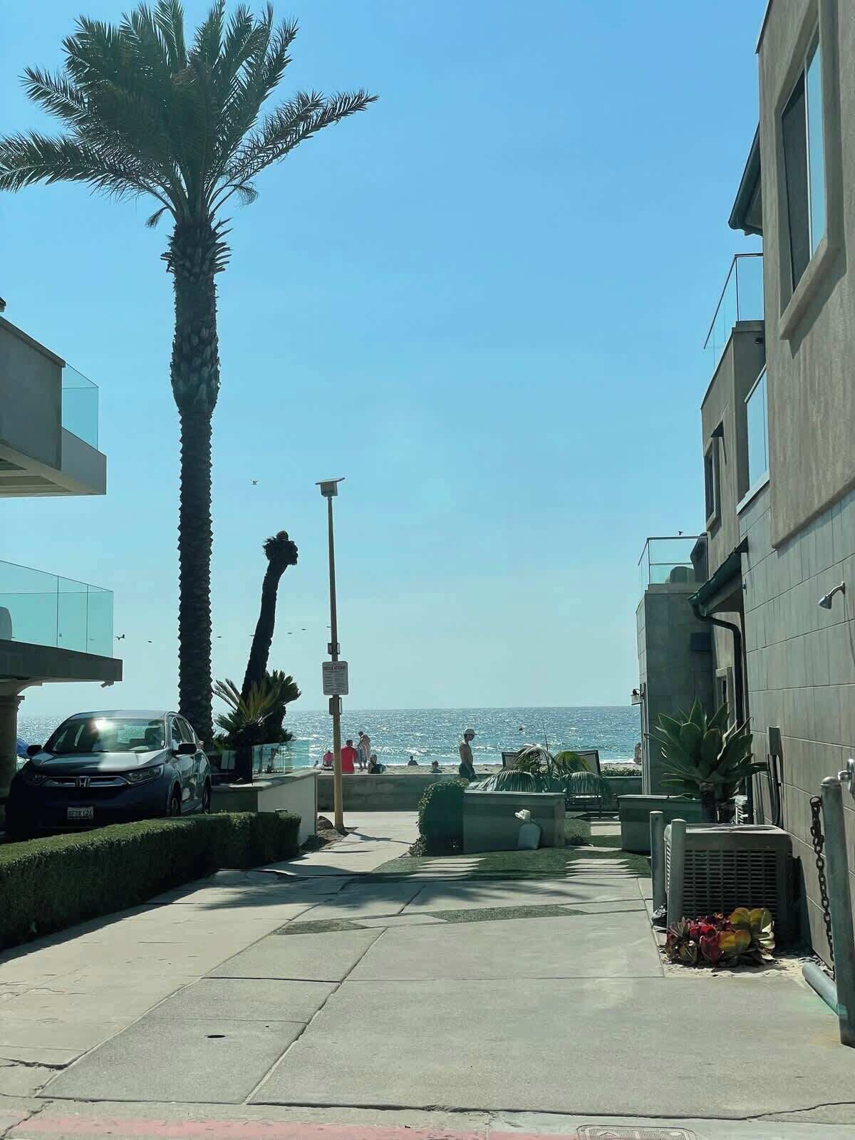 Een kuststraat met een palmboom, een geparkeerde auto en een gebouw met een balkon dat uitkijkt over het strand.