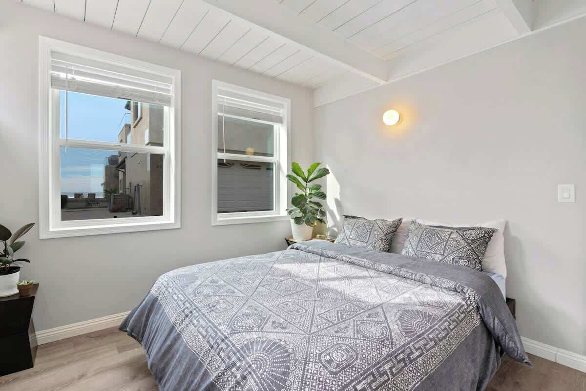A bedroom with a bed, two windows, a potted plant, and a wall-mounted light fixture.