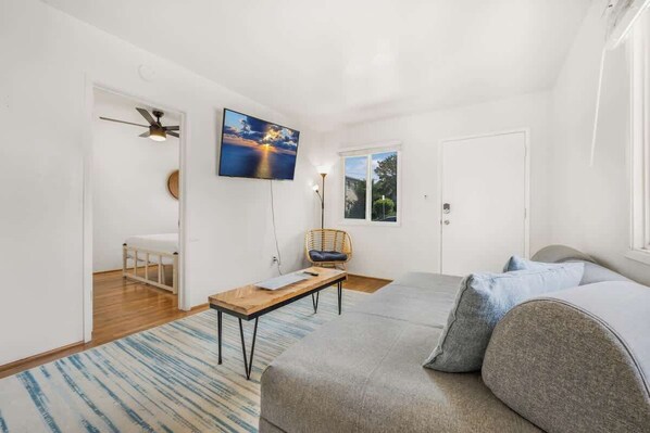 A modern, well-lit room with a large bed, a small table, a chair, and a TV mounted on the wall. - Fresh Pacific Beach Surf Pad BBQ/Fire Pit-Sleeps 4 (San Diego)