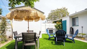 A backyard with a dining table, chairs, a thatched umbrella, and a grill.
