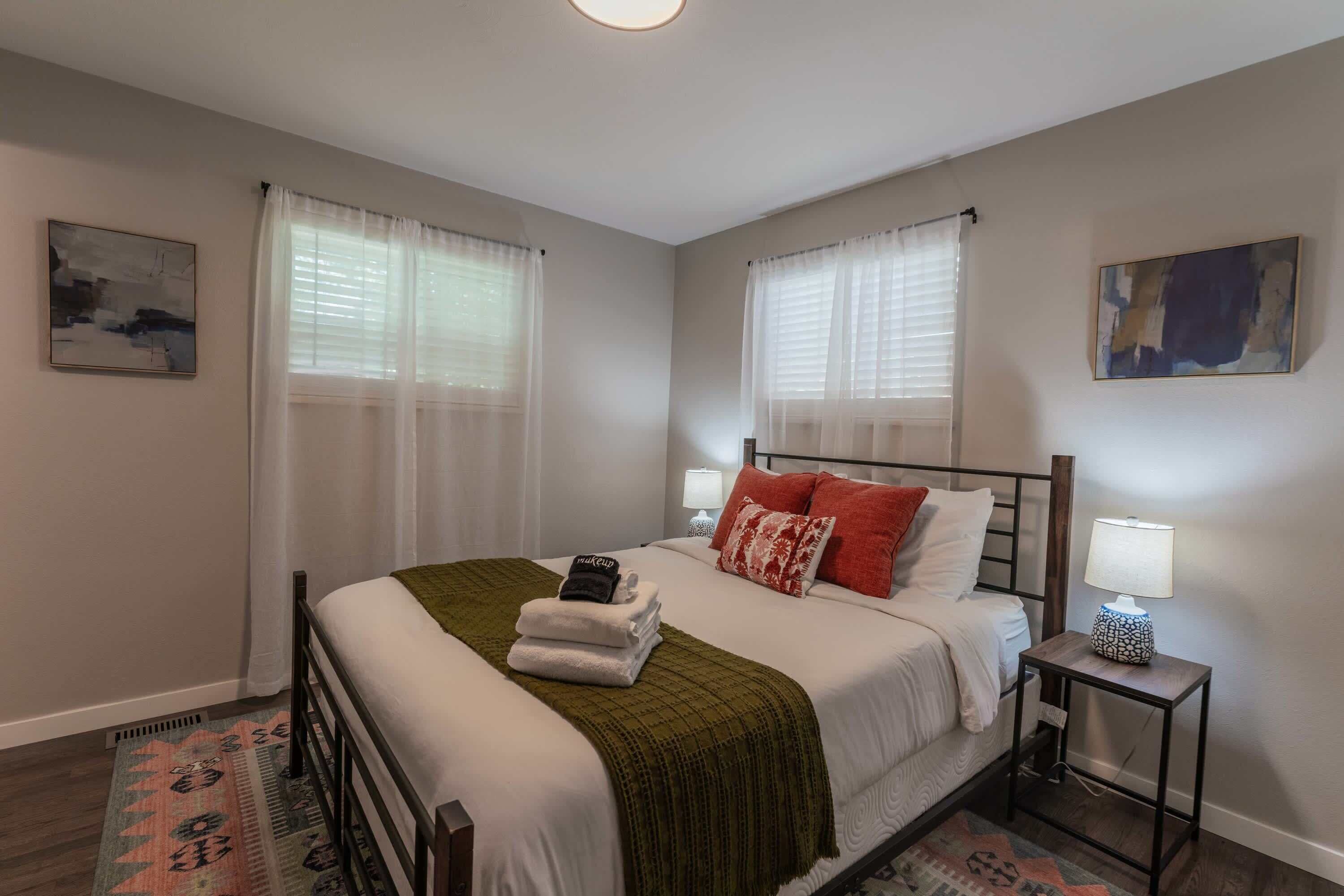A neatly made bed with a green blanket, white pillows, and a red pillow. There is a small bedside table with a lamp and a wall-mounted painting.