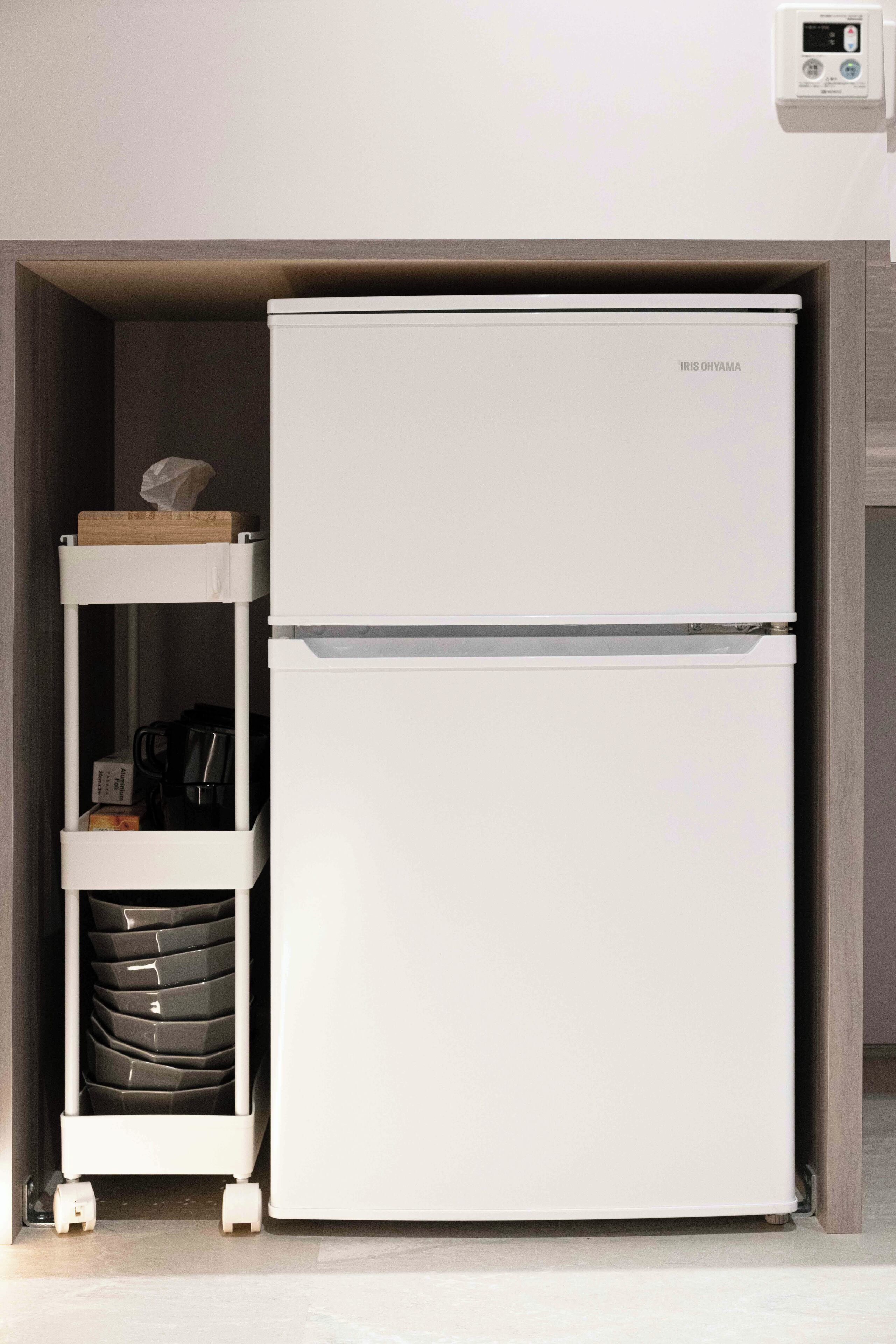 A compact, white refrigerator with a digital display, placed in a narrow space with a small shelf unit beside it.