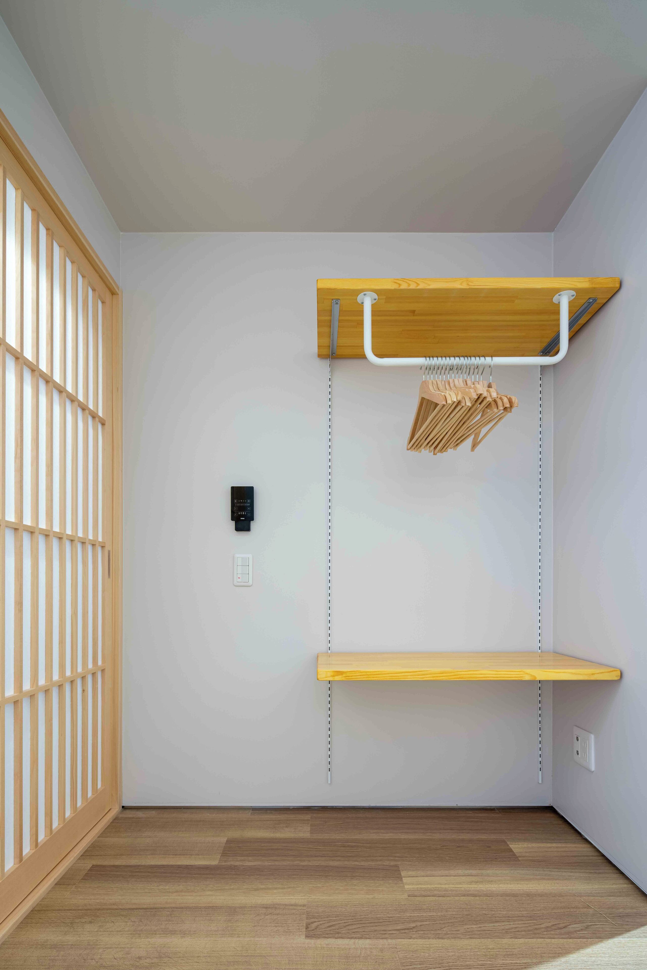 A room with a wooden floor, a low wooden shelf, a wall-mounted shelf with a hanging rack, a power outlet, and a sliding door with vertical blinds.