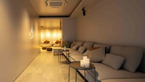 A modern spa room with a large beige sofa, a small round table with candles, and a wall-mounted massage chair. - KUTSUROGI SAUNA and STAY (Tokyo)