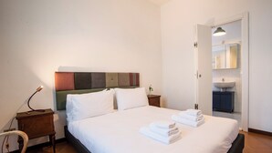 Una camera d'albergo con un letto, un comodino, una lampada e un bagno con lavandino e specchio.