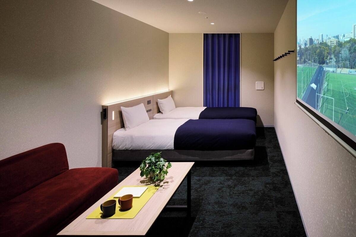 Quarto de hotel com cama, sofá vermelho, mesa pequena com planta e duas chávenas, e uma janela ampla com vista para um campo desportivo e edifícios.