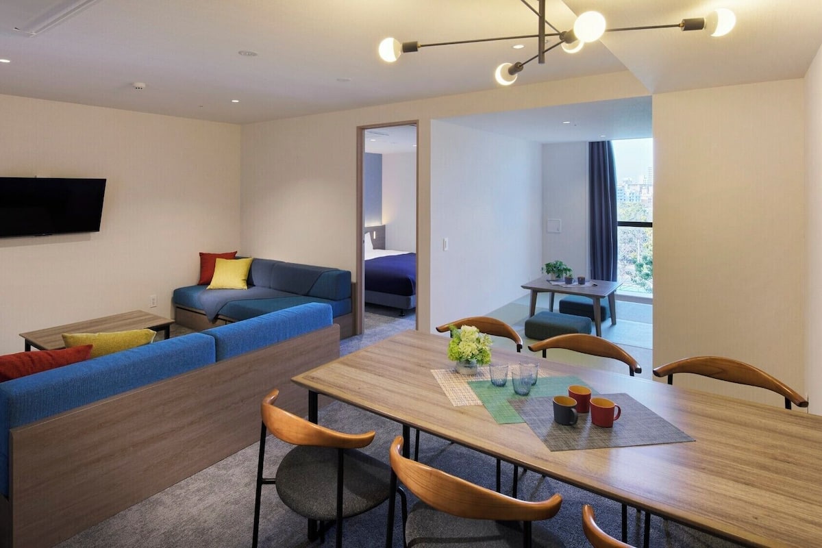 Quarto de hotel moderno com sofá, mesa de jantar com cadeiras, TV de tela plana e vista da cidade por uma janela grande.