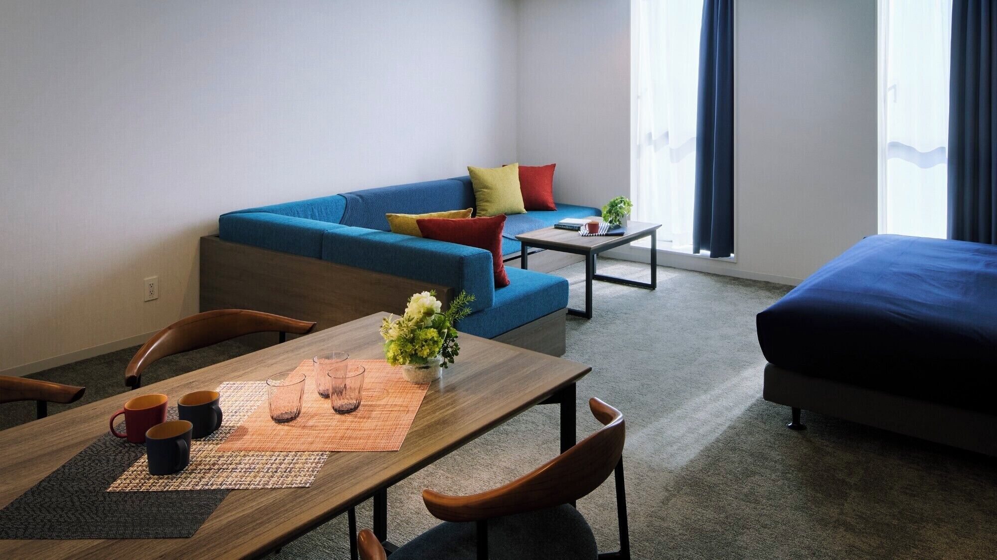 Una sala de estar moderna con un sofá azul, una mesa de comedor de madera con sillas, una pequeña mesa auxiliar y una cama.