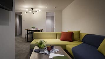 Un salon moderne avec un canapé vert, un canapé bleu, une table basse en bois et, au fond, un coin repas.