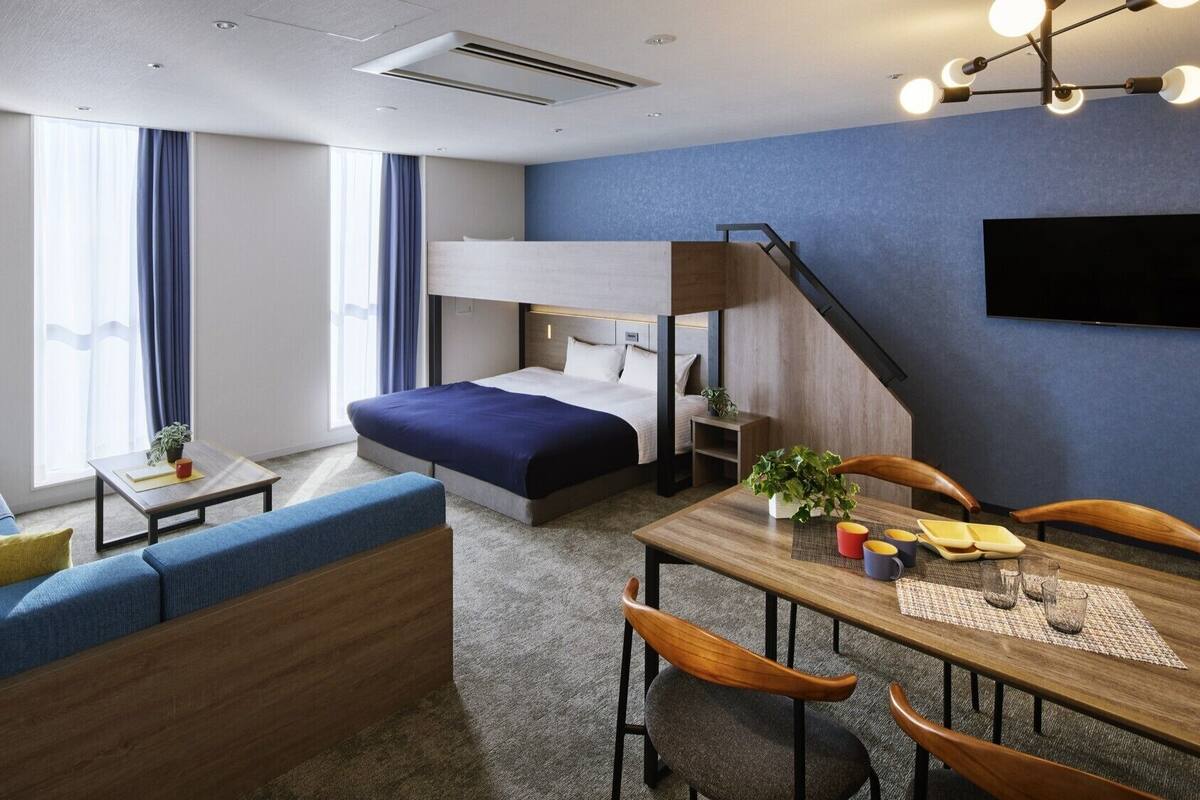 Quarto de hotel moderno com uma beliche, um sofá, uma mesa de jantar com cadeiras e uma TV fixada na parede.