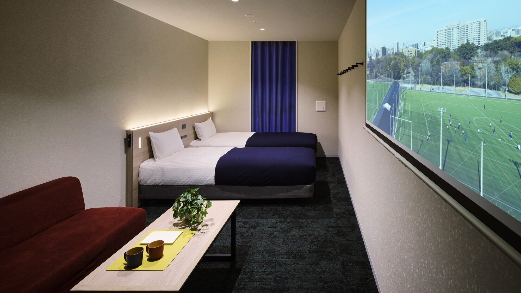 Hotelkamer met een bed, een bank, een kleine tafel met een plant en een raam met uitzicht op een sportveld.
