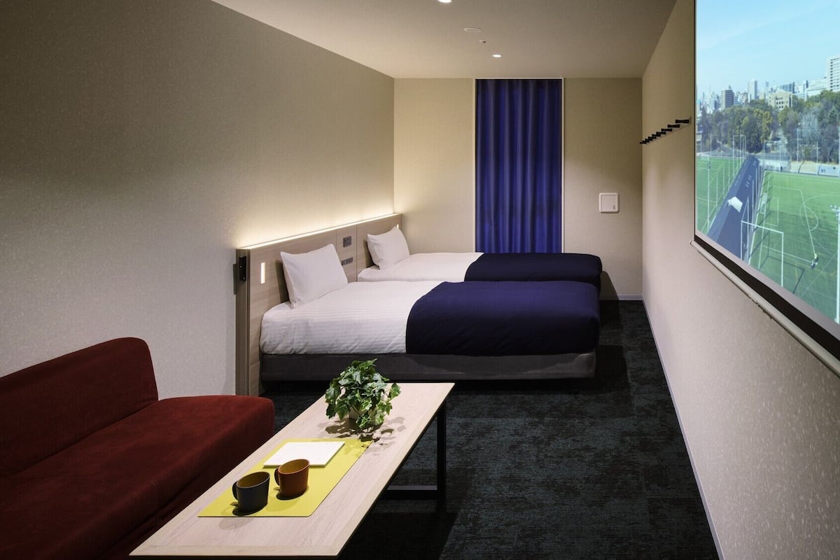 Habitación de hotel con cama, sofá, una mesita con una planta y ventana con vistas a un campo deportivo.