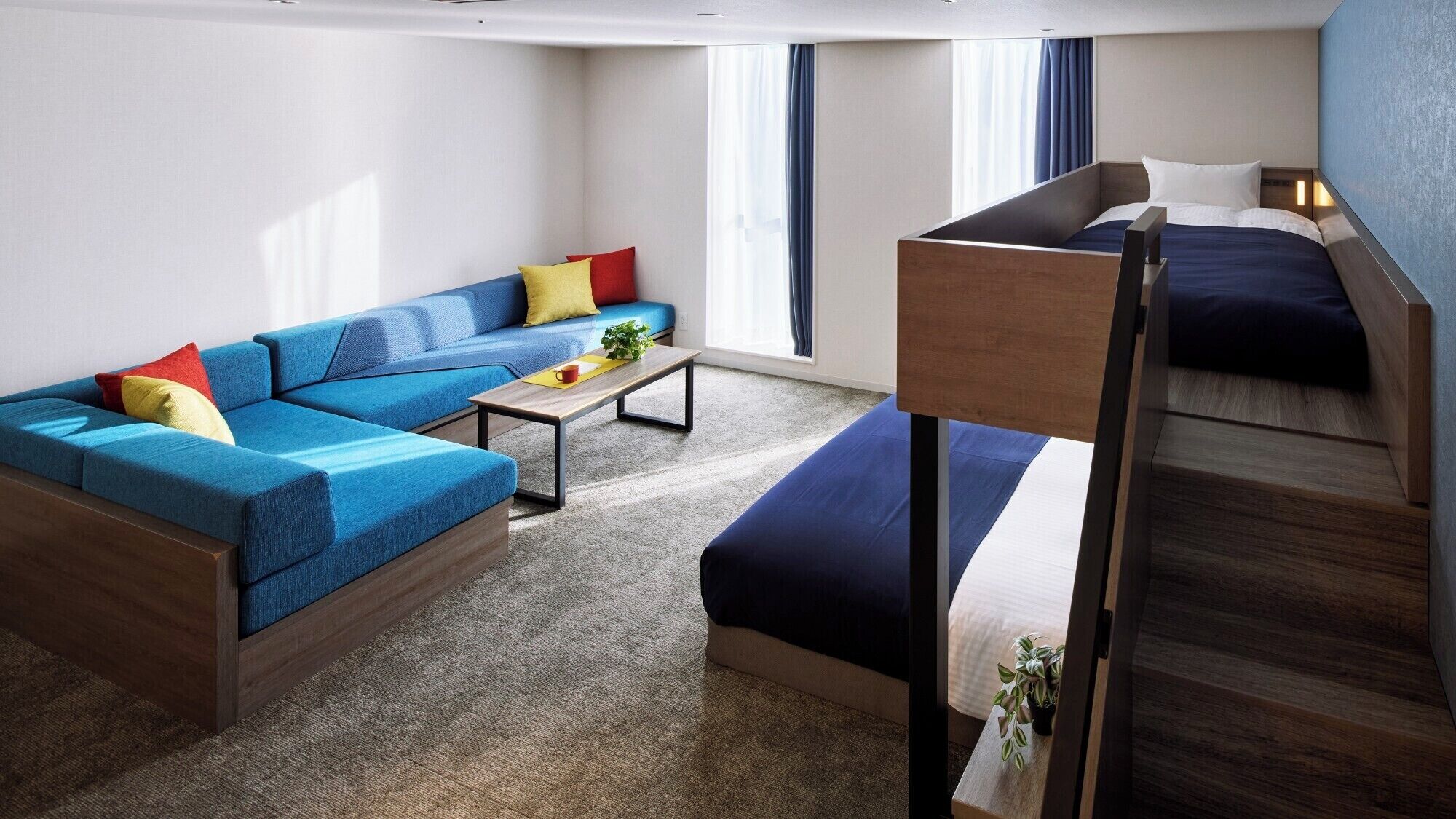 Una habitación de hotel compacta con sofá, una mesita, una litera y un ventanal grande con cortinas.
