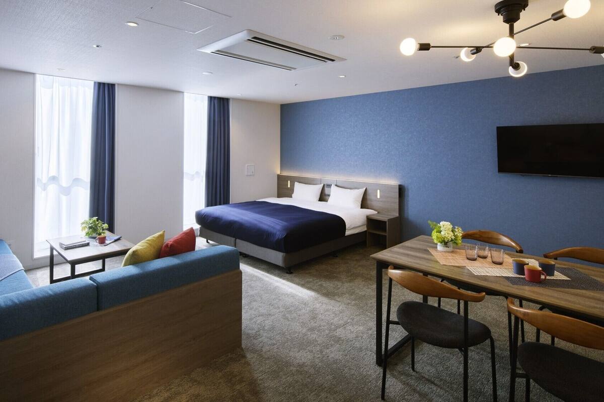 Quarto de hotel moderno com uma cama grande, um sofá, uma mesa de jantar com cadeiras e uma televisão de tela plana.