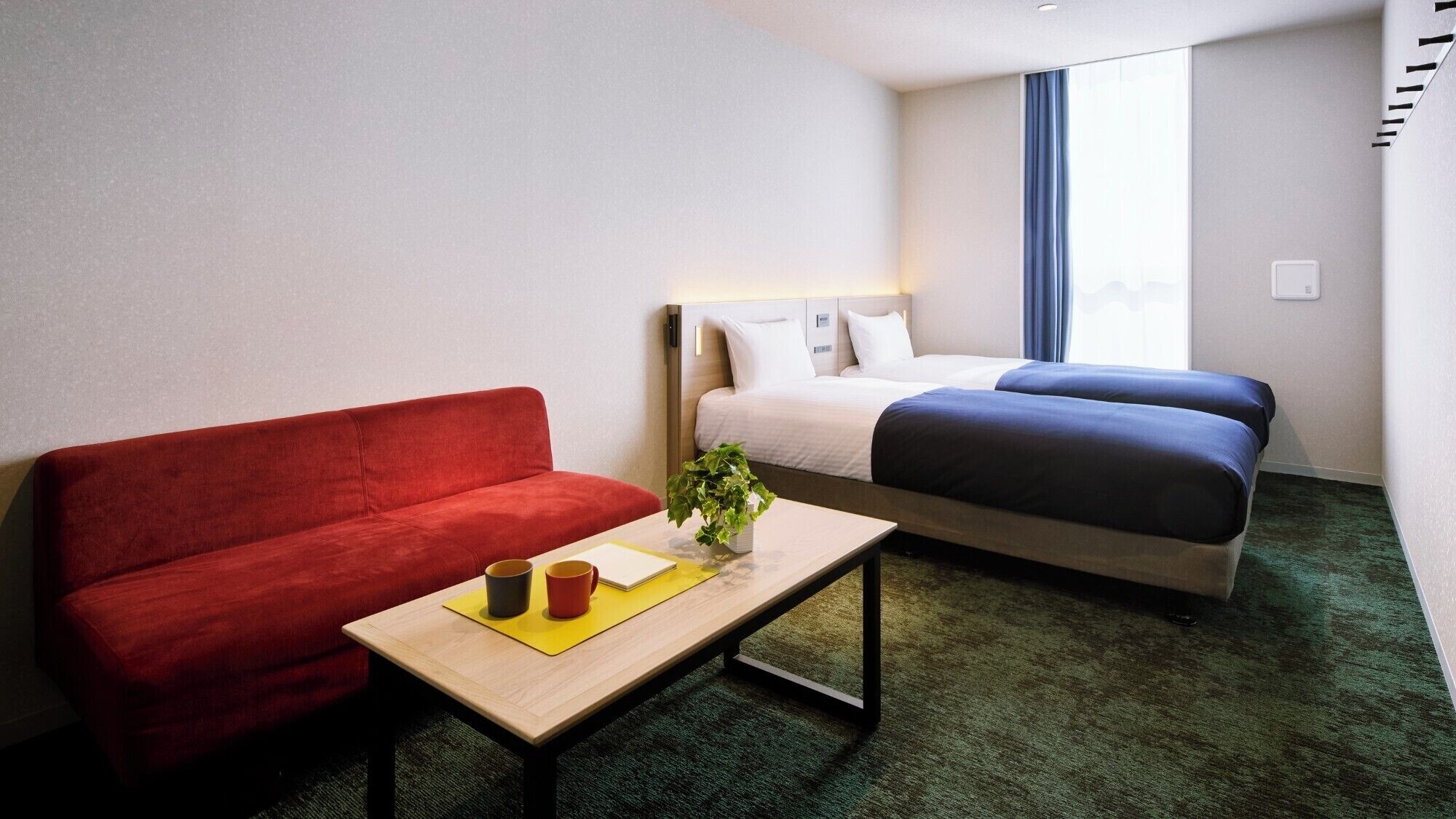 Habitació d'hotel amb un sofà vermell, una taula de fusta amb una planta, un llit amb roba de llit blava i una catifa verda.