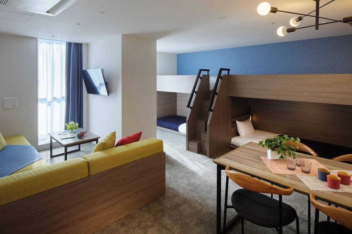 Quarto de hotel moderno com sofá, mesa de jantar com cadeiras, televisão montada na parede e beliche com escada.