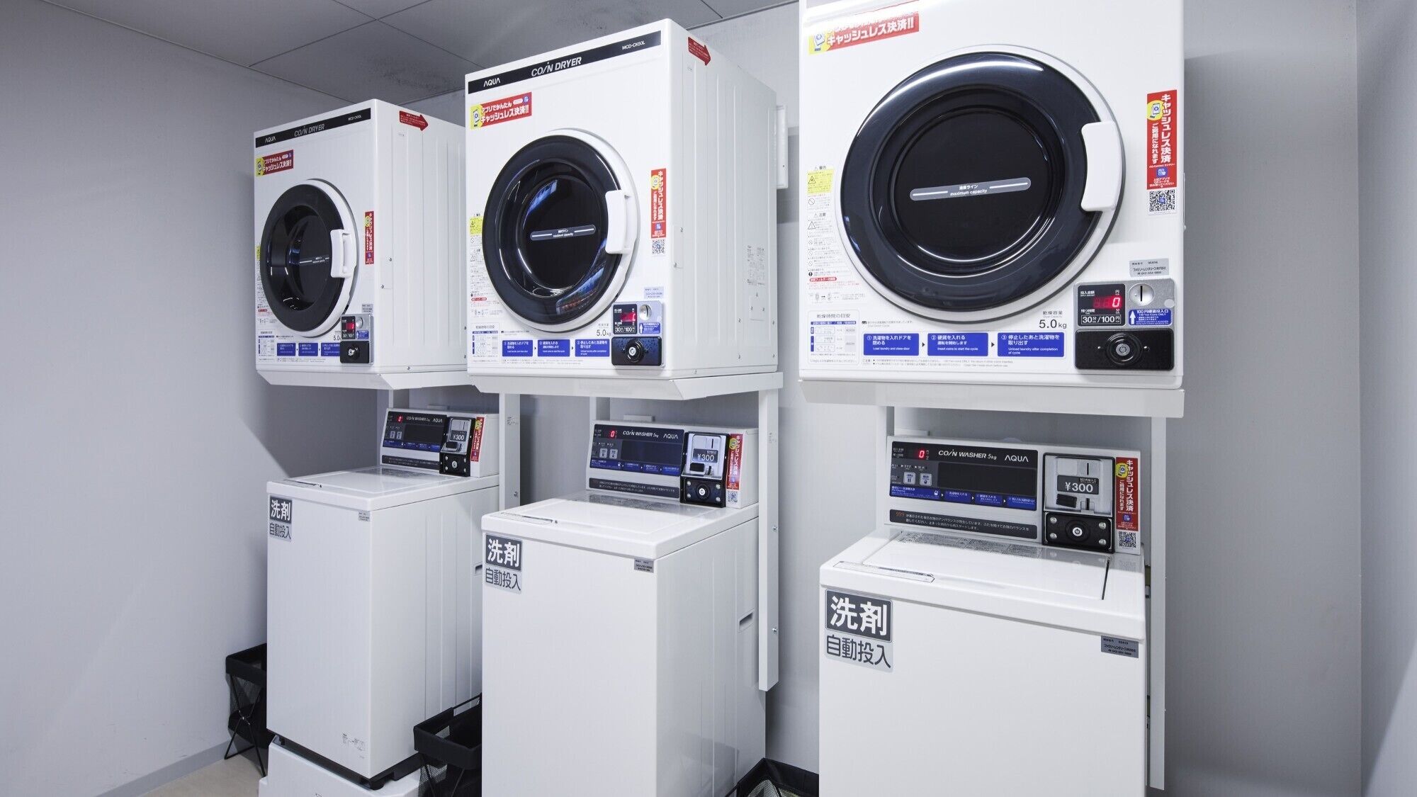 A row of white, front-loading washing machines with digital displays and control panels, arranged in a commercial setting.