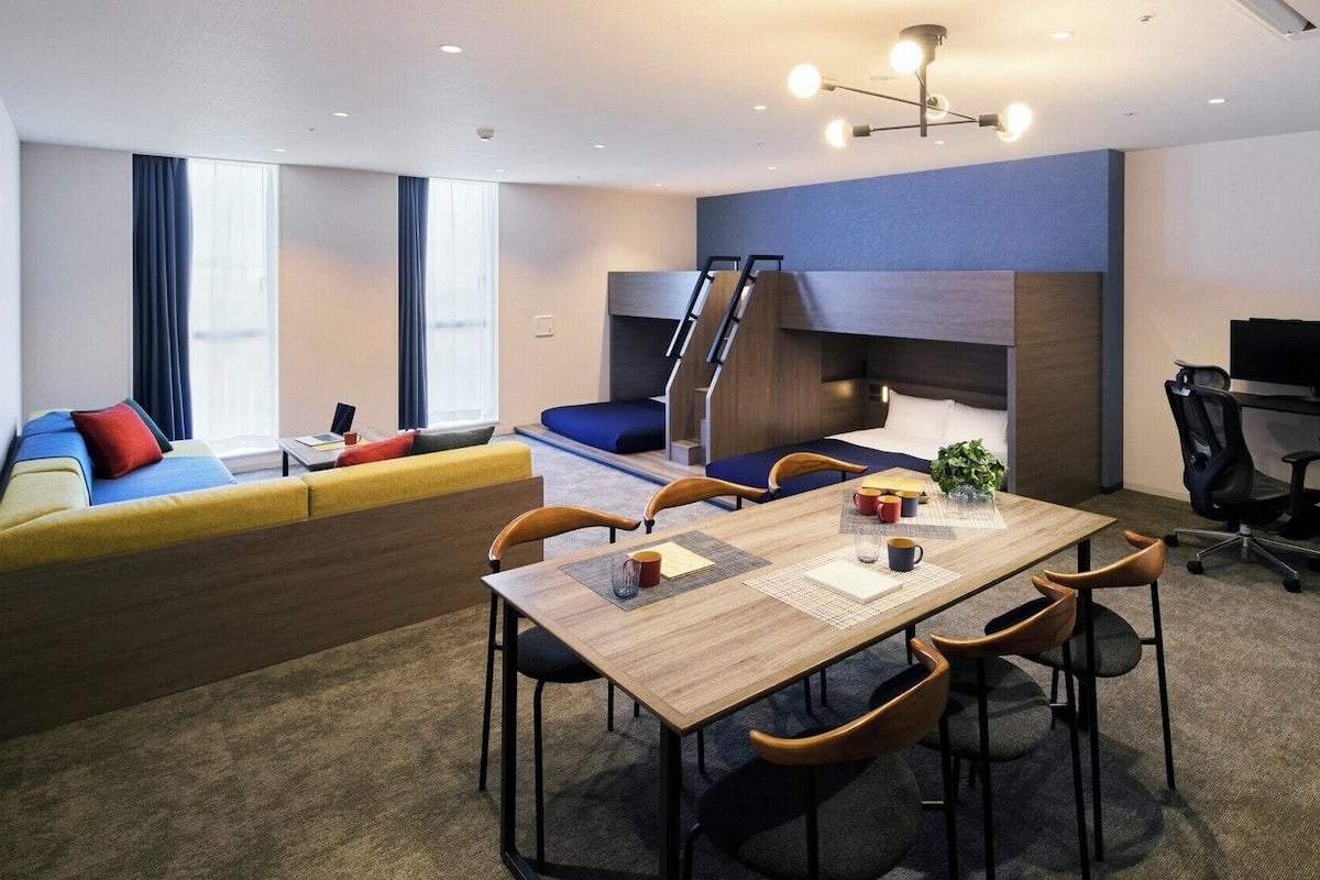 Quarto de hotel moderno com área de refeições, sofá com almofadas coloridas, escrivaninha com computador e uma grande janela.
