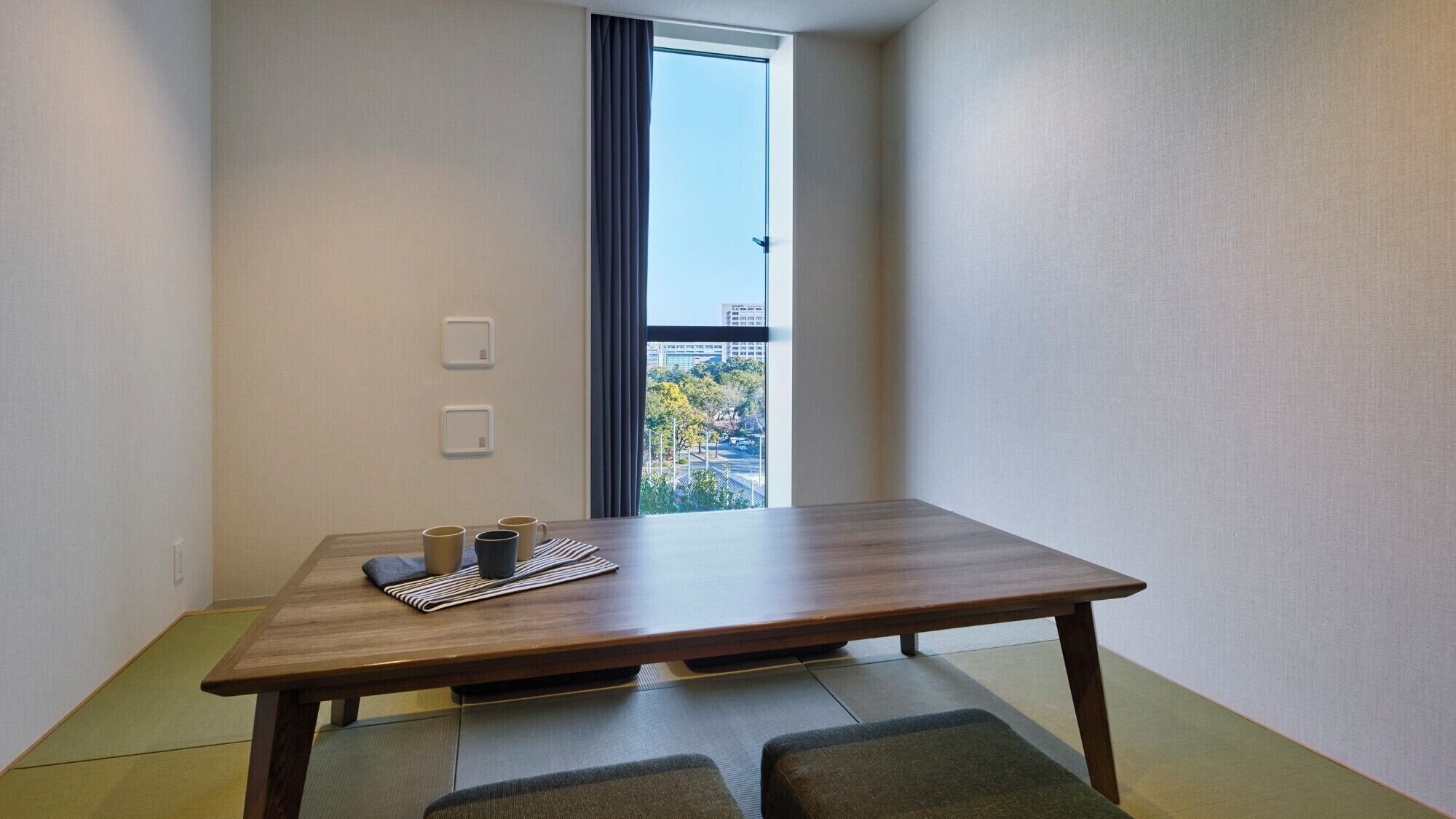 A room with a wooden table, two chairs, a window with a view of trees and buildings, and two wall-mounted light switches.