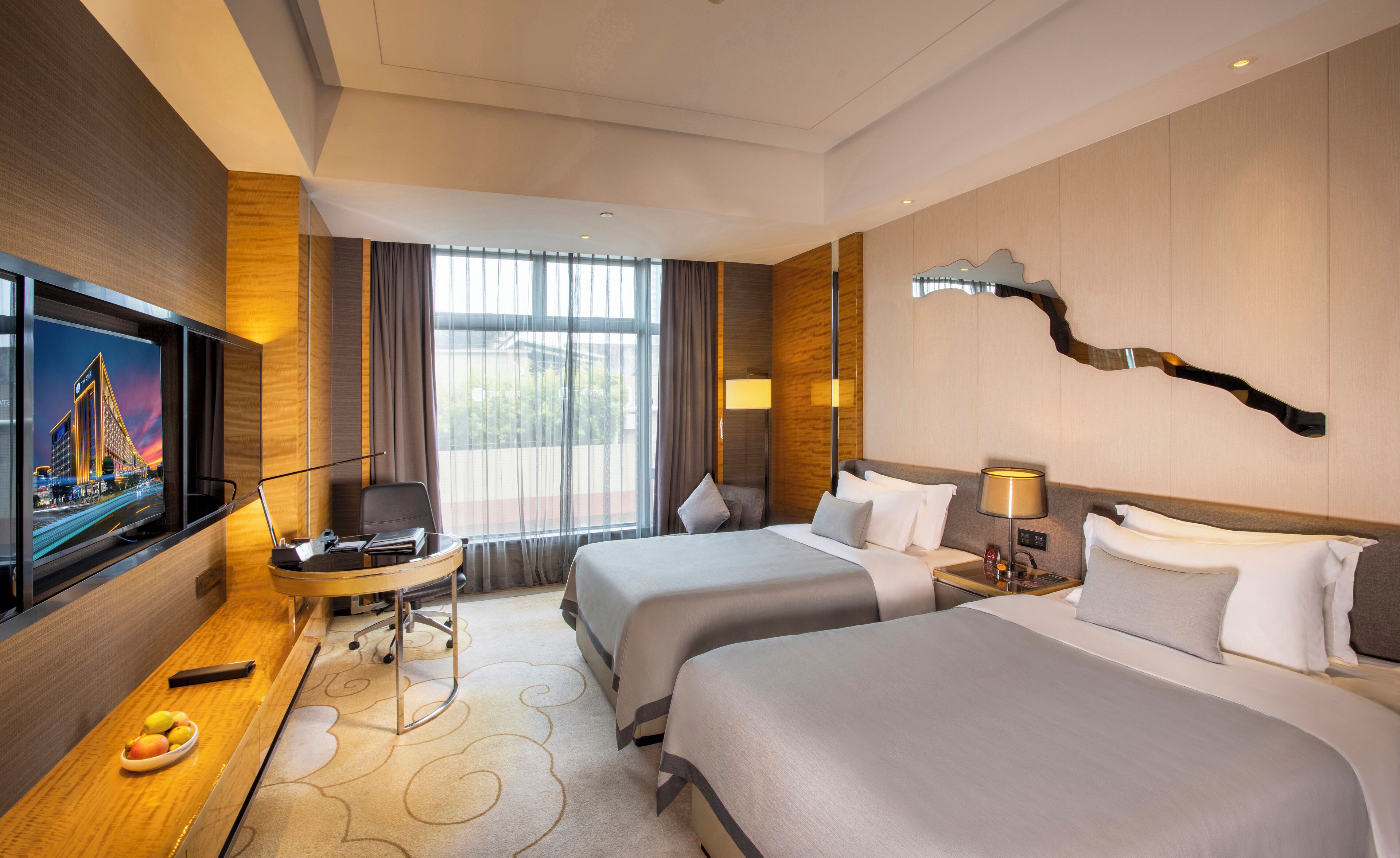 Quarto de hotel com duas camas, uma escrivaninha com cadeira, televisão, abajur e uma cesta de frutas.