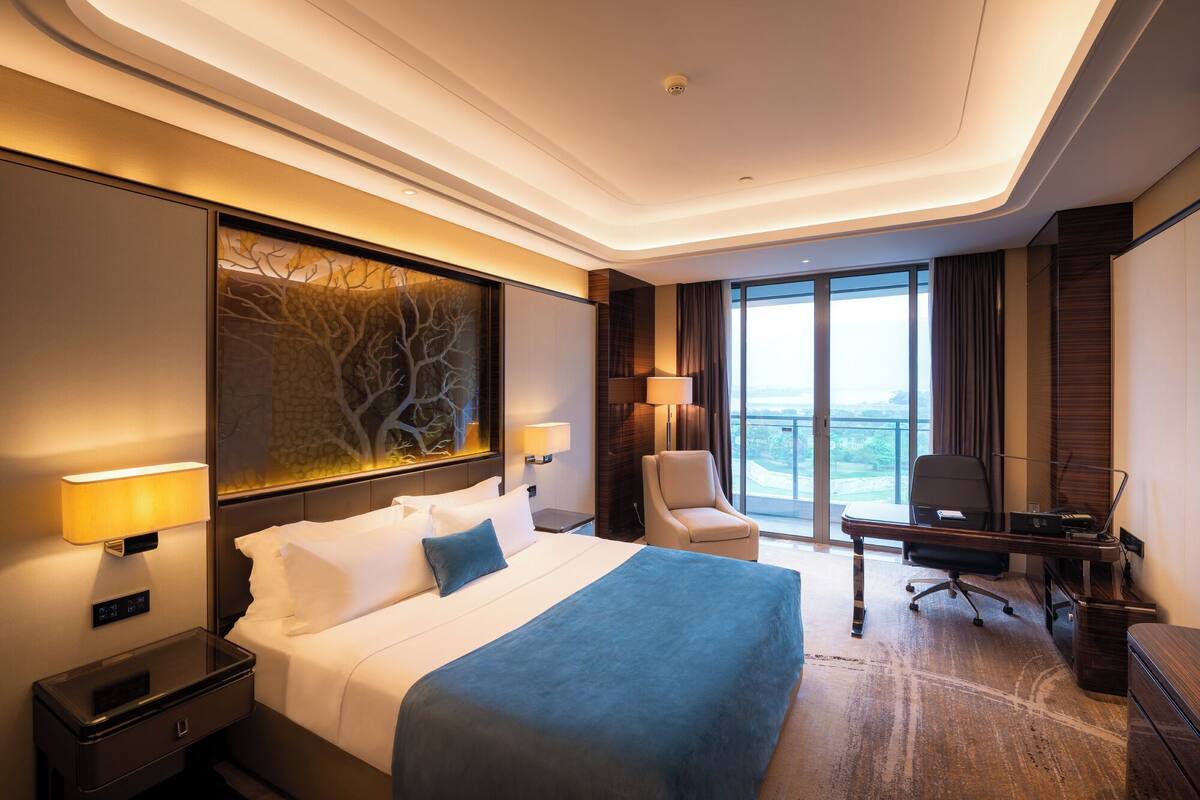 A modern hotel room with a large bed, a desk, a chair, and a view of the city.