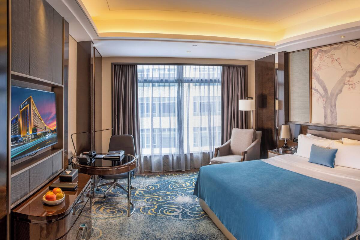A hotel room with a large bed, a desk with a computer, a chair, a TV, and a large window with curtains.