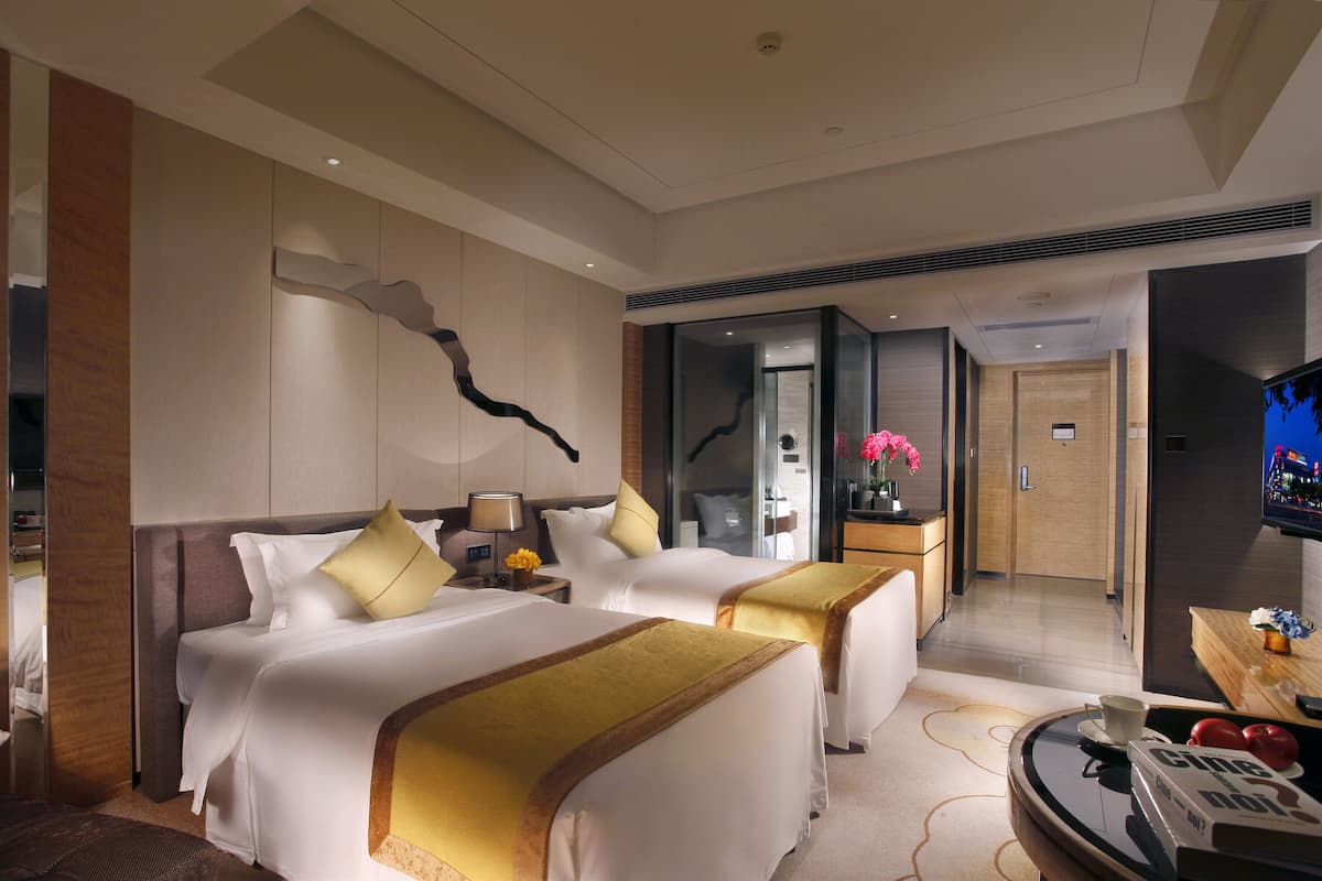 A hotel room with two beds, a TV, a desk with a chair, and a bathroom visible through a glass door.