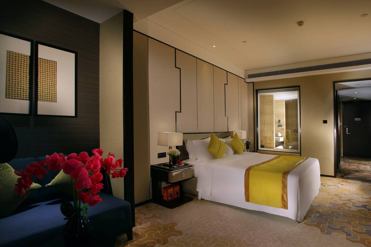 A hotel room with a bed, a nightstand, a lamp, a vase with flowers, and a sofa.